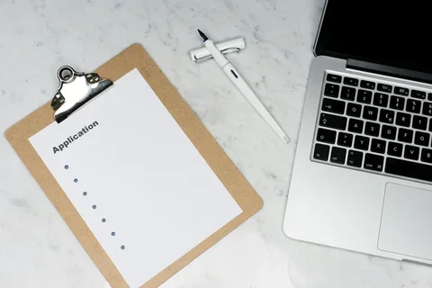 Clipboard with a blank application form beside a laptop and pen on a marble desk.