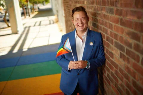 Man smiling with white shirt and blue jacket holding the LGBTQAI+ flag