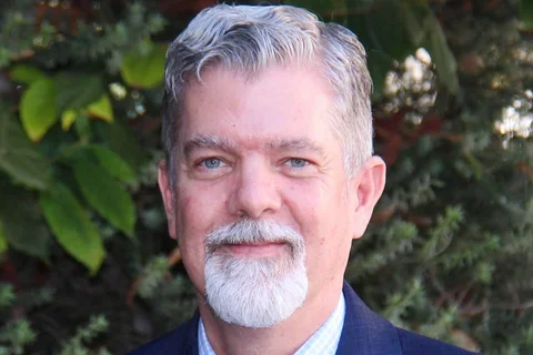 Gray-haired bearded man in a blue suit with an ID badge, standing outdoors.