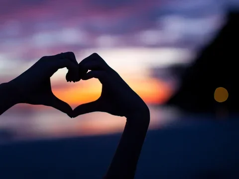 Two hands creating a heart in front of a sunset