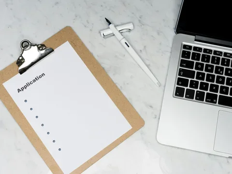 Clipboard with a blank application form beside a laptop and pen on a marble desk.