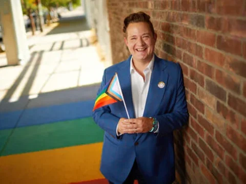 Man smiling with white shirt and blue jacket holding the LGBTQAI+ flag