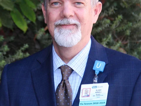 Gray-haired bearded man in a blue suit with an ID badge, standing outdoors.